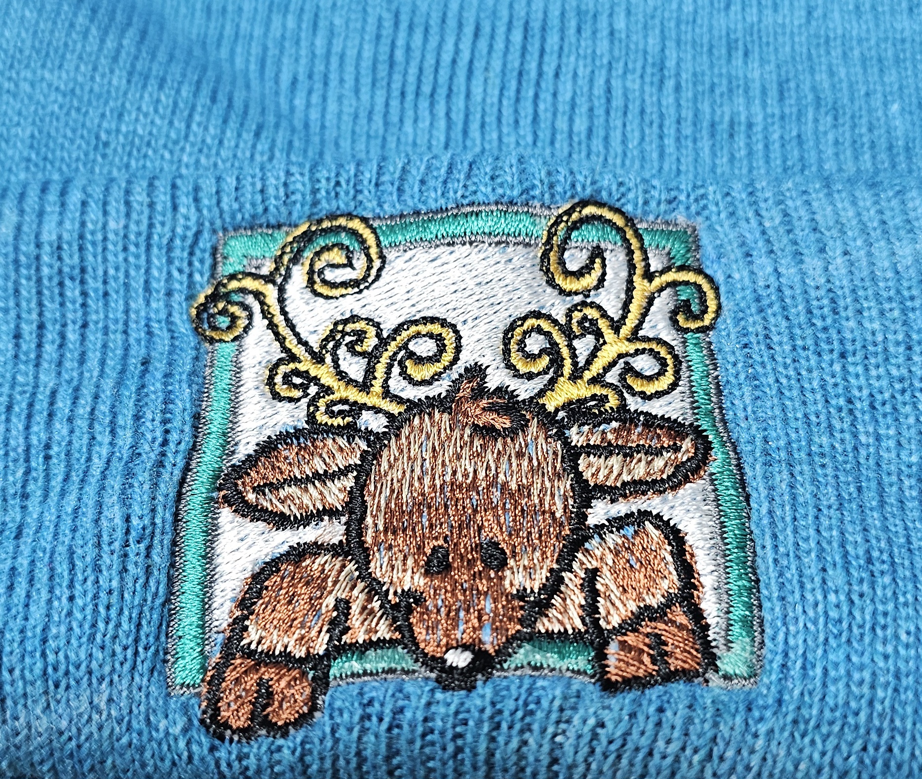 Reindeer peeking over a framed patch embroidered on a blue beanie