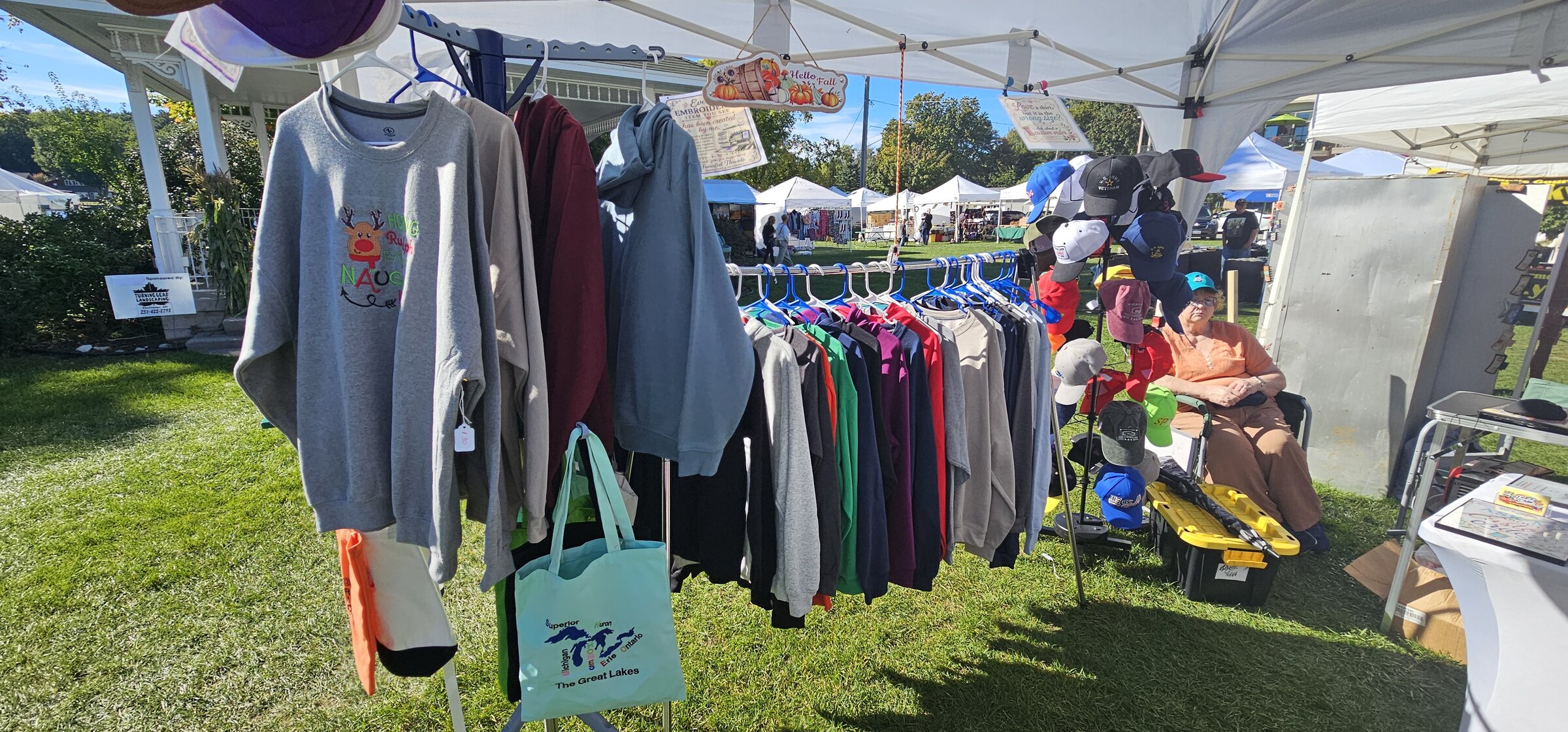 Racks of embroidered sweatshirts, hats, and tote bags at an outdoor Fanciful Threads craft fair booth
