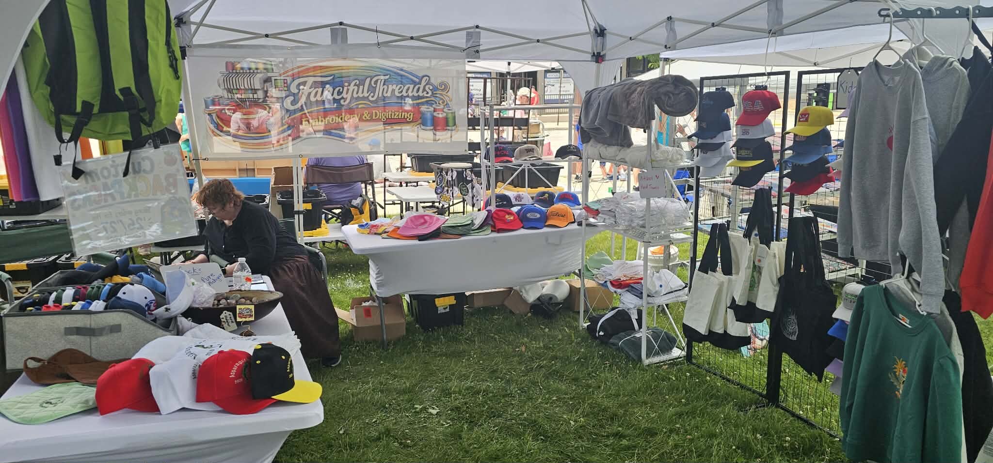 Fanciful Threads booth displaying embroidered hats, apparel, bags, and accessories at an outdoor craft fair