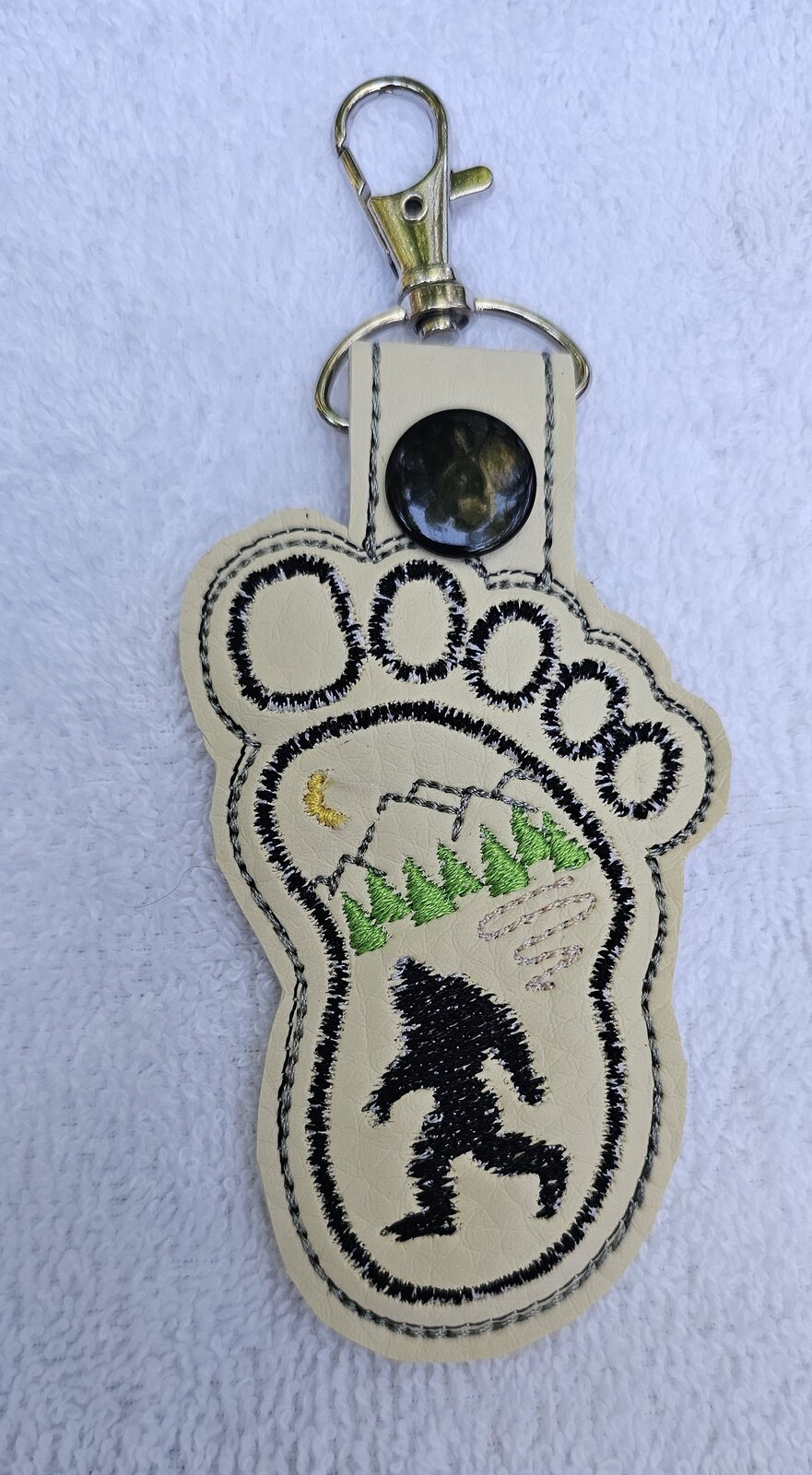 Cream footprint key fob embroidered with Bigfoot, mountains, and pine trees