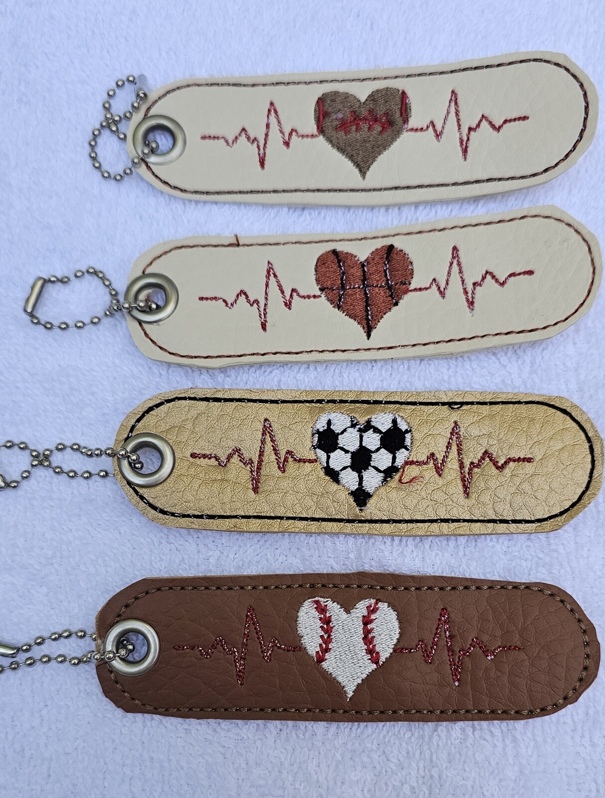 Four tag key fobs embroidered with heartbeat lines and football, basketball, soccer, and baseball hearts