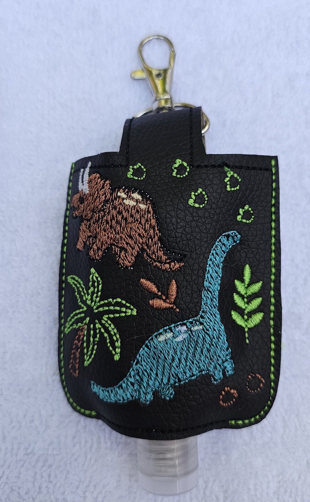 Black hand sanitizer holder embroidered with dinosaurs, palm trees, and footprints
